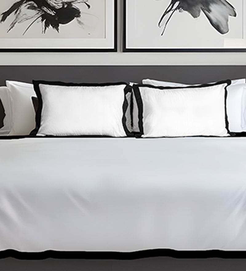 White bedding set with black trim on a bed, framed artwork in the background.
