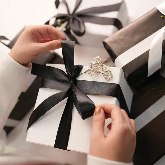 Person adjusting a black ribbon on a gift box with another gift in the background.